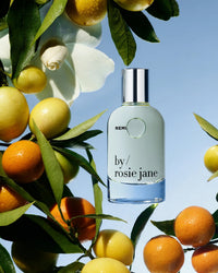 A bottle of Rosie Jane Eau De Parfume in Remi is placed among green leaves, citrus fruits, and a white flower, all set on a blue background.