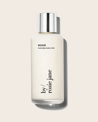 A bottle of Rosie Jane Rosie Everyday Body Milk, featuring a silver cap and creamy white lotion with soft white musk notes, is displayed against a light beige background.