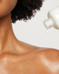 A close-up of a person’s bare shoulder and neck with curly hair; a white bottle dispenses Rosie Jane Rosie Everyday Body Milk, hydrating their skin. The soft scent of white musk lingers against the plain, light background.