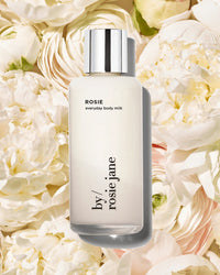 A bottle of Rosie Everyday Body Milk by Rosie Jane with a silver cap is shown against a background of white peony, rose flowers, and subtle notes of white musk.