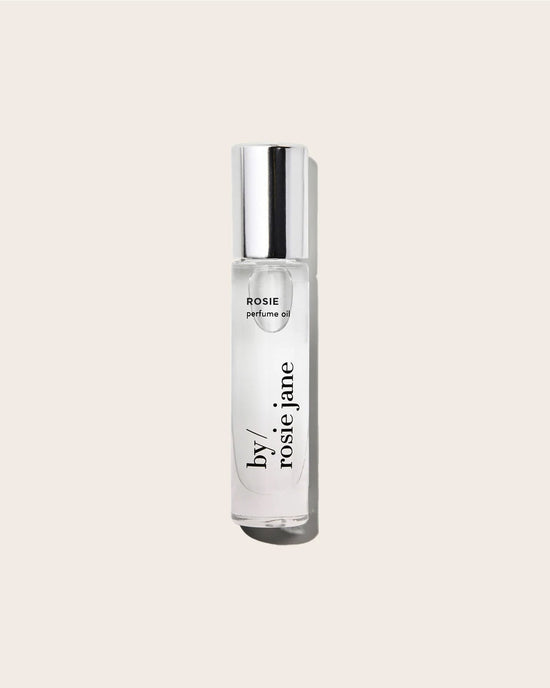 The Perfume Oil in Rosie by Rosie Jane is a clear glass bottle with a silver cap, shown against a beige background. This fragrance offers a delicate skin scent with soft notes of white musk.