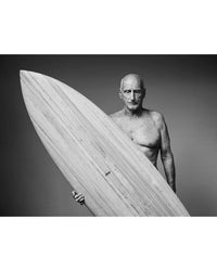 A shirtless older man with a bald head holds a large surfboard upright against a plain backdrop. Featured in "Surf Tribe" by Simon & Schuster, this classic black-and-white photo captures the essence of surf culture.