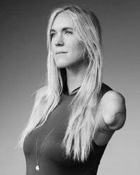 A black-and-white portrait of a woman with long blonde hair and one arm amputated above the elbow, wearing a sleeveless top and necklace, as featured in "Surf Tribe" by Simon & Schuster—reflecting resilience and thoughtful introspection.