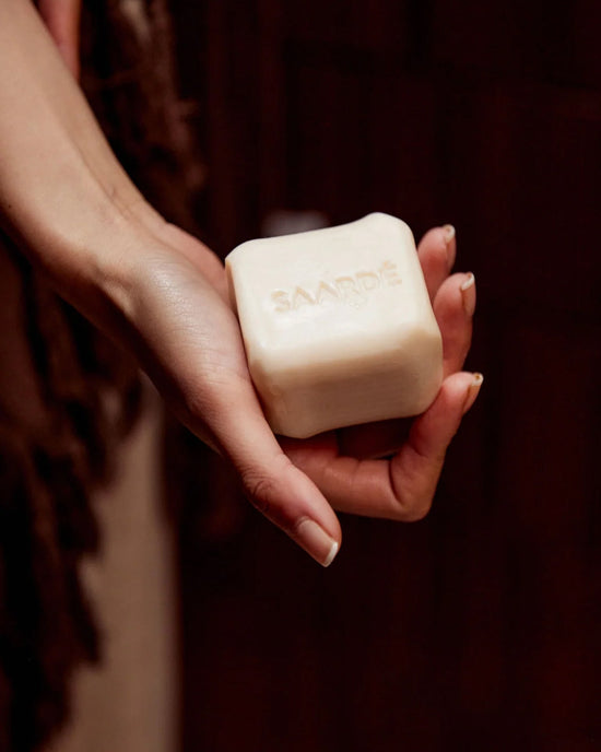 A hand holds a rectangular Saarde Olive Oil Bar Soap in Medina, embossed with the brand name, against a dark background.