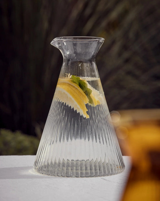 The Saarde Kairos Angular Carafe in Clear, filled with water, lemon slices, and mint leaves, rests on a white surface against a softly blurred background.