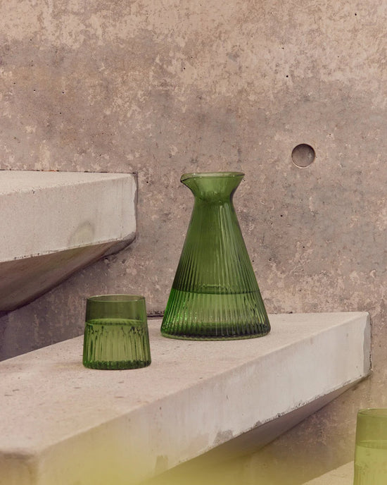 The Saarde Kairos Medium Water Glass in Green sits on angled concrete shelves with a matching carafe, set against a textured concrete wall.
