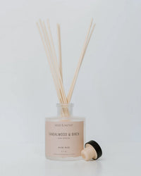 The Near & Native Room Diffuser in Sandlewood & Birch features a glass bottle with reed sticks and a black wooden cap, providing continuous fragrance, all displayed against a plain white background.