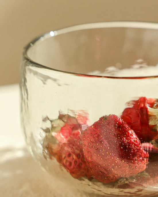 A clear hand-blown Pebbled Bowl from The Collective holds ripe strawberries with green stems, displayed on a light-colored surface.