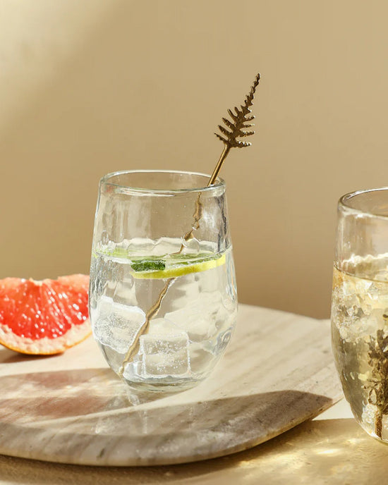 A refreshing drink with ice, cucumber, and a grapefruit wedge is elegantly served in The Collectives Pebbled Small Glasses – Set of 4, featuring a stylish hammered design.