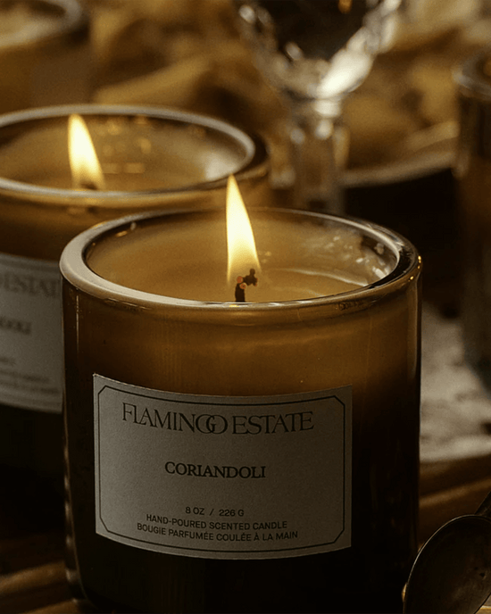 A lit Coriandoli Candle by Flamingo Estate, crafted with vegetable wax and enclosed in a glass jar, glows warmly while another candle and soft background objects enhance the cozy atmosphere.