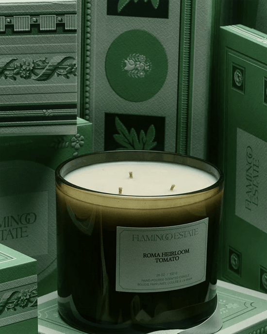 The Tomato XL Candle by Flamingo Estate, crafted with vegetable wax, rests among ornately designed books and packaging featuring embossed patterns and botanical motifs.