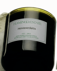 A close-up of Flamingo Estates Prinsesstarta Candle, an 8 oz almond marzipan hand-poured candle in a dark glass jar, shown resting on a white surface.