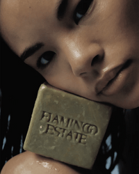 A person with wet hair holds a square Green Goddess Bar Soap by Flamingo Estate, embossed with the brand name, close to their face.