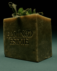 The Green Goddess Bar Soap by Flamingo Estate is a dark green, rectangular bar embossed with the brand's name and topped with leafy sprigs, capturing its bold essence against a deep background.