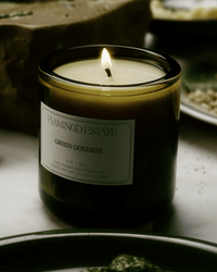 The Flamingo Estate Green Goddess Candle, hand poured from vegetable wax, glows in a dark green glass jar on a table with softly blurred plates and cozy, warm lighting.