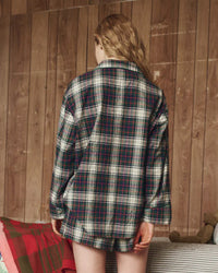 A person with wavy blonde hair, seen from behind, wears the Great. Sleep's Flannel Pajama Top in Pine Lodge Plaid with matching shorts, standing in a rustic room with wood-paneled walls and a nearby blanket.