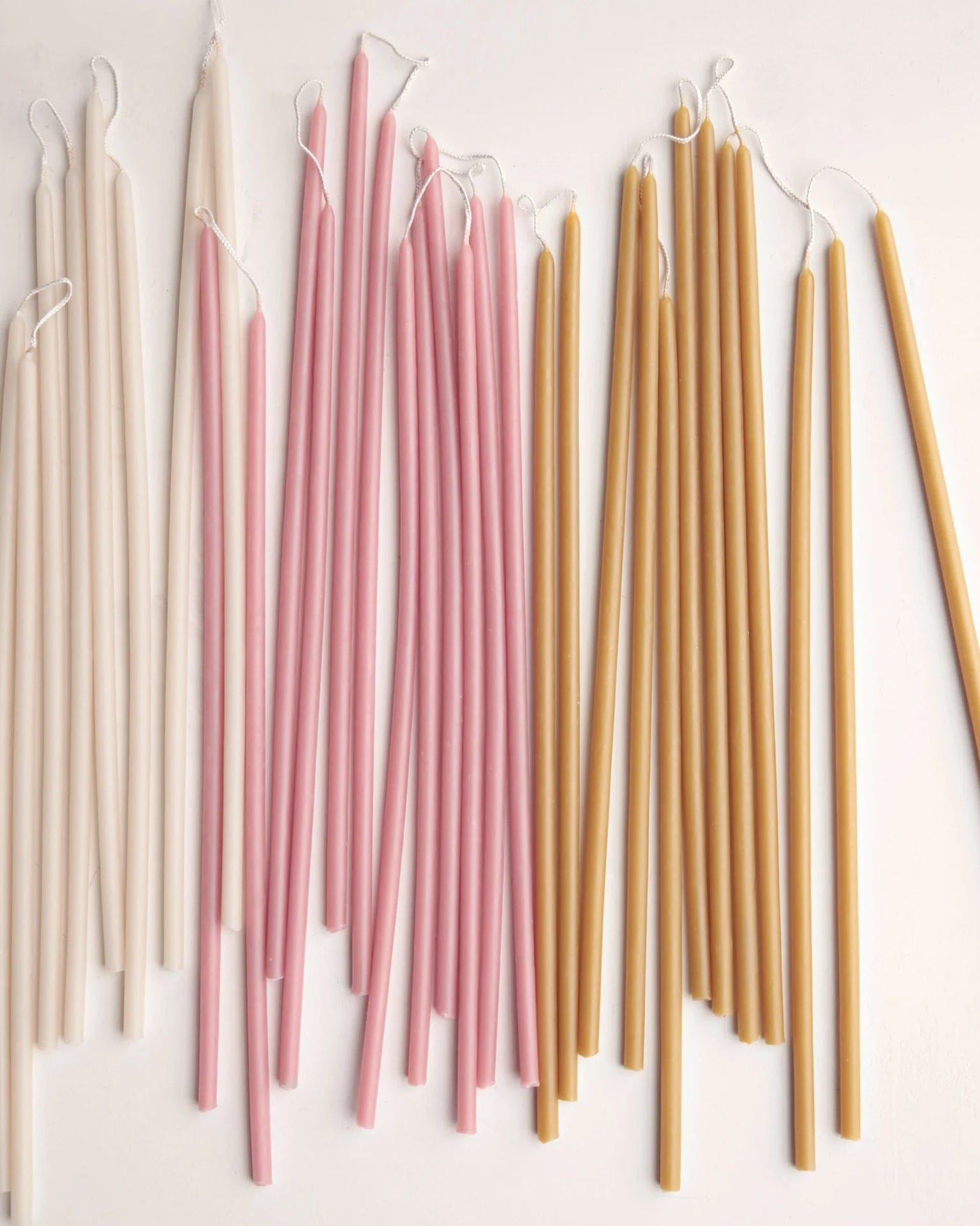 Beeswax Long Stem Candles by The Floral Society are displayed in rows on a light surface. These tall, slender candles in off-white, pink, and mustard yellow are grouped by color, each featuring unburned wicks.