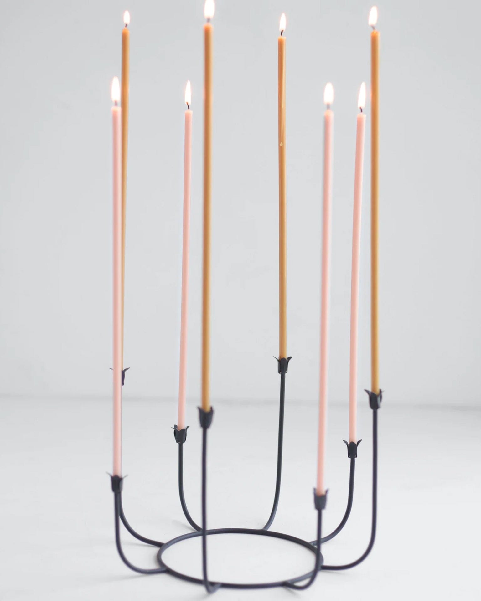 A black metal candle holder displays seven tall Beeswax Long Stem Candles by The Floral Society—four pink and three gold—arranged in a circle against a plain white background.