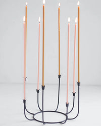 A black metal candle holder displays seven tall Beeswax Long Stem Candles by The Floral Society—four pink and three gold—arranged in a circle against a plain white background.