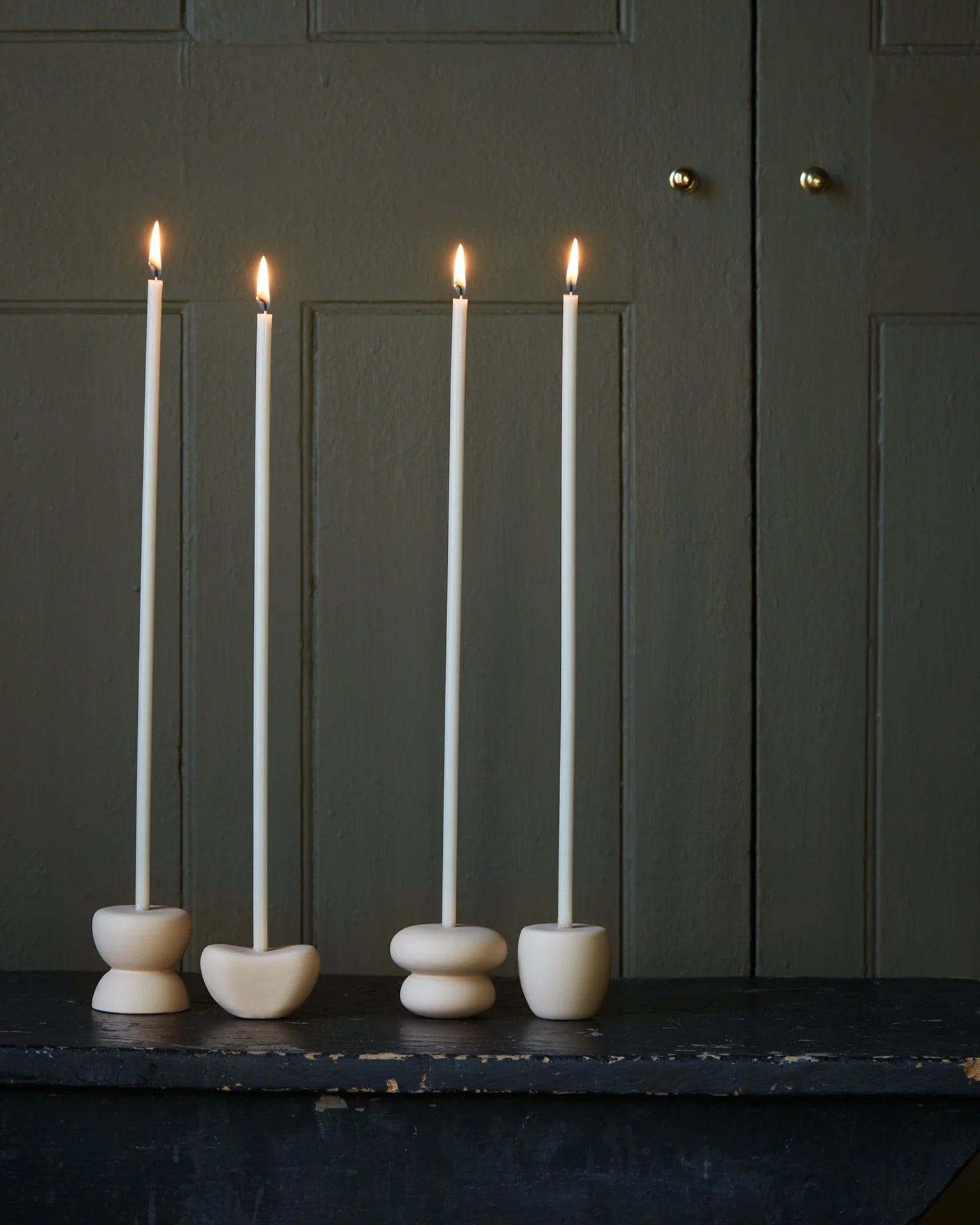 Four lit Beeswax Long Stem Candles by The Floral Society, in minimalist round holders, sit on a dark wooden surface against a muted green paneled background.
