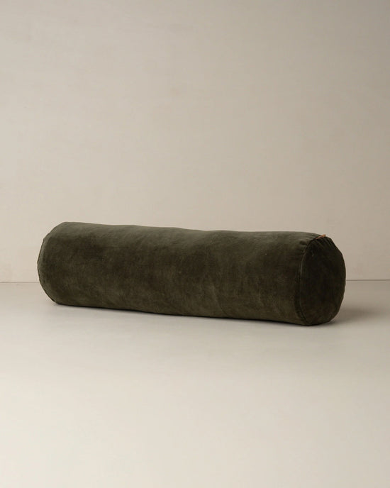 The Saarde Velvet Structured Bolster in Dark Olive rests on a plain, light surface against a neutral wall.