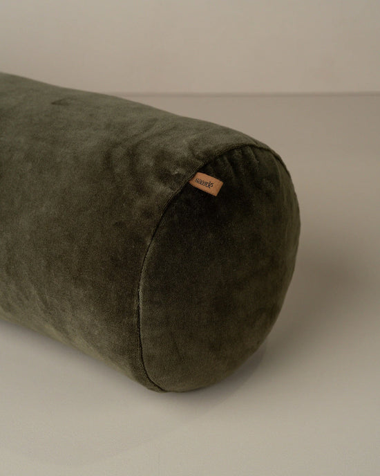 The Saarde Velvet Structured Bolster in Dark Olive, a cylindrical cushion with a small fabric tag, is shown resting on a light surface.