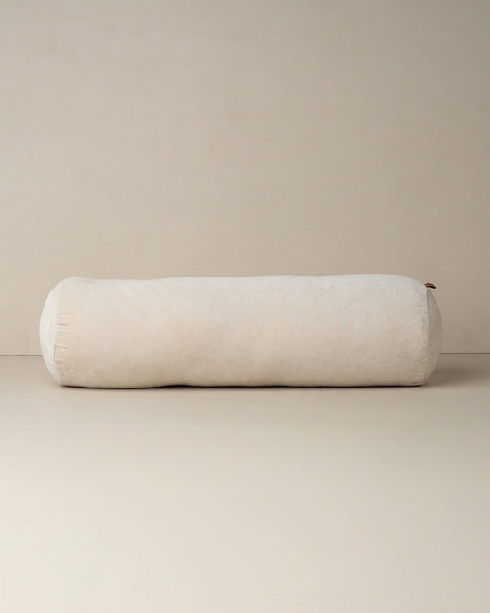 The Saarde Velvet Structured Bolster in Clay, featuring a large cylindrical shape, rests on a neutral beige surface with a matching backdrop.