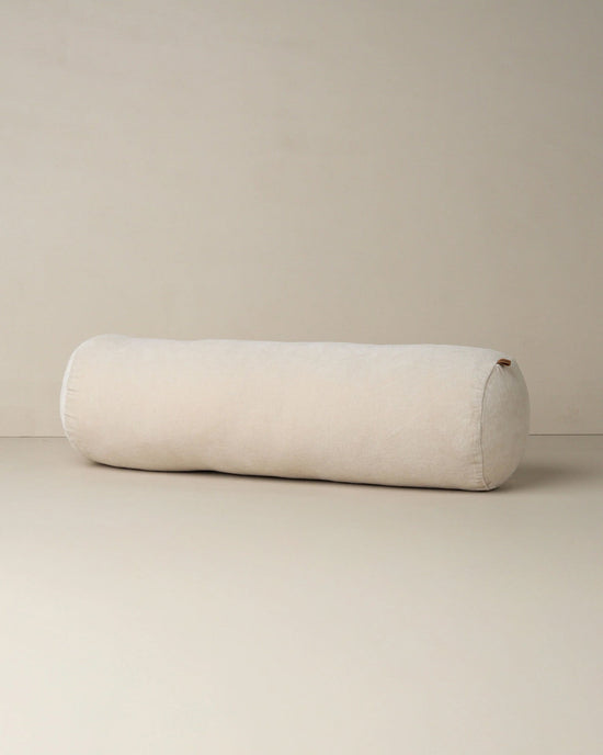 The Saarde Velvet Structured Bolster in Clay, a long cylindrical cushion, rests on a light-colored floor against a plain, light wall.
