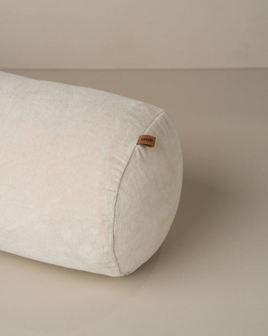 The Saarde Velvet Structured Bolster in Clay is a cylindrical off-white cushion with a small brown fabric tag on one end, displayed on a smooth, neutral surface.