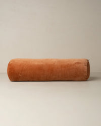The Saarde Velvet Structured Bolster in Terracotta rests on a neutral floor beside a plain wall, showcasing its large cylindrical cotton velvet design.