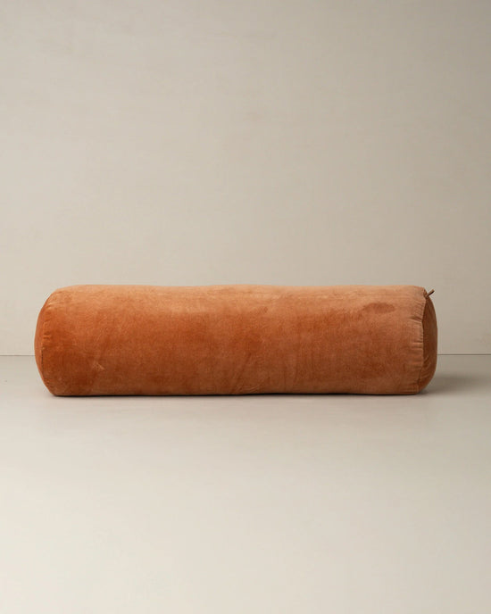 The Saarde Velvet Structured Bolster in Terracotta rests on a neutral floor beside a plain wall, showcasing its large cylindrical cotton velvet design.