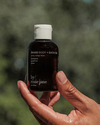 A hand holds Rosie Jane's Wake Body + Bath Oil, a small dark bottle featuring skin-hydrating ingredients such as eucalyptus, grapefruit, and lemon, with a blurred green background adding to its refreshing vibe.