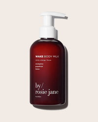 A dark amber pump bottle labeled "Wake Body Milk" by Rosie Jane, features hydrating key ingredients—eucalyptus, grapefruit, and lemon—and is shown against a light beige background.