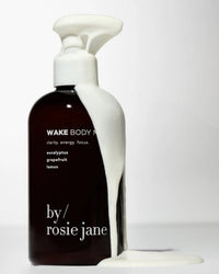 A dark bottle labeled Wake Body Milk by Rosie Jane features hydrating white body milk overflowing from its pump and dripping down the side, pooling at the base, set against a plain white background.