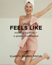 A smiling woman wrapped in a pink towel jumps joyfully. Text reads: Rosie Jane Wake Body Milk—feels like morning sunshine and a splash of cold water. Hydrate skin. CLARITY. ENERGY. FOCUS.