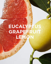 A close-up of a grapefruit slice and a lemon with green leaves on white. Overlaid text: Rosie Jane Wake Body + Bath Oil—Eucalyptus, Grapefruit, Lemon, and hydrating kakadu plum.