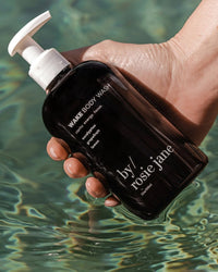 A hand holds a dark pump bottle labeled Wake Body Wash by Rosie Jane above clear, sunlit pool water.