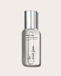 A 3 fl oz clear bottle of Rosie Jane’s White Tee Body + Hair Perfume Mist with a silver cap and cotton blossom notes, shown against a light beige background.