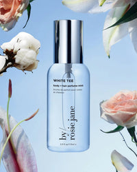 A clear bottle of Rosie Jane White Tee Body + Hair Perfume Mist is centered on a sky-blue background with soft clouds, surrounded by cotton blossom, roses, and white flowers.