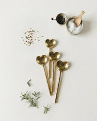 Four Casa Amarosa Heart Shaped Brass Cocktail Spoons (set of 4) are displayed on a white surface with fresh rosemary, spices, a mini wooden spoon, salt bowl, and peppercorns—an elegant choice for stylish barware lovers.