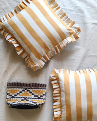 Two Casa Amarosa Golden Yellow Ruffle 16x16in pillows with yellow and white stripes rest on a light fabric surface. A small rectangular pouch with a brown, white, and yellow geometric pattern lies below them.
