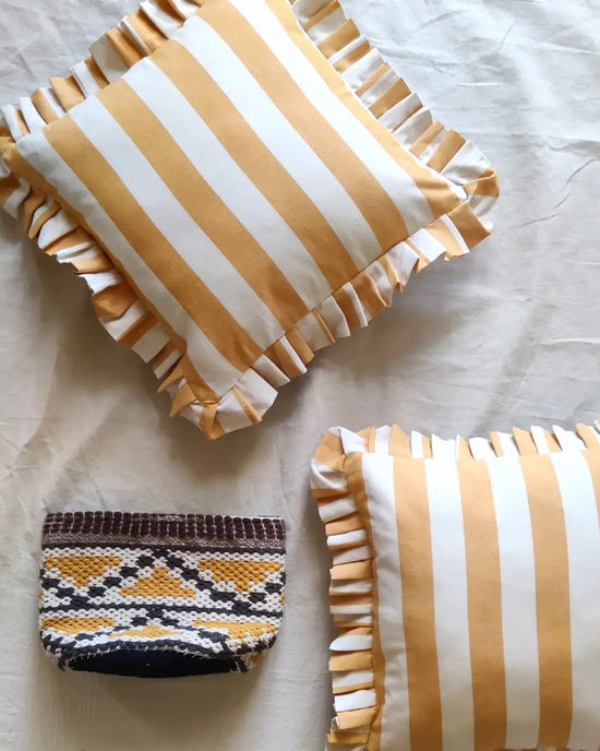 Two Casa Amarosa Golden Yellow Ruffle 16x16in pillows with yellow and white stripes rest on a light fabric surface. A small rectangular pouch with a brown, white, and yellow geometric pattern lies below them.