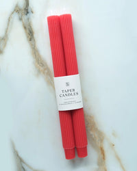 Two Cherry Taper Candles | Fluted by BOTANICA, hand-dyed in red with a white paper label, are placed diagonally on a white marble surface featuring brown and gray veins.