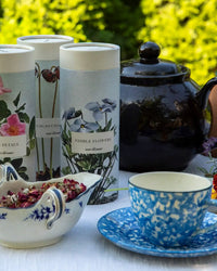 Enjoy a serene outdoor tea with The Quiet Botanist's Edible Flower Blend, paired with a blue-patterned teacup, dark teapot, and floral canisters—ideal for adding edible flowers to tea or cakes. Lush greenery sets the scene.