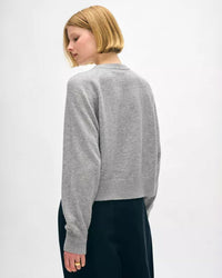 A woman with short blonde hair wears the White + Warren Cashmere Easy Sweatshirt in Grey Heather and dark pants, standing with her back to the camera and glancing over her shoulder against a plain white background.