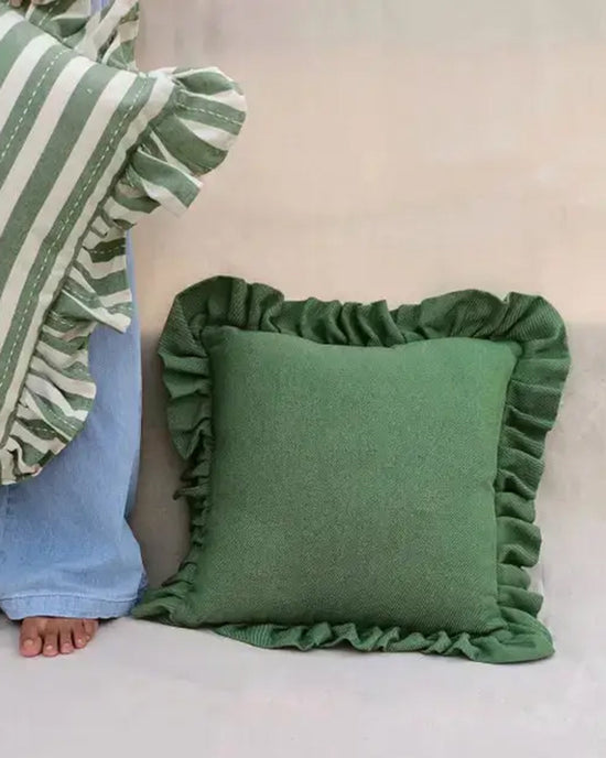 A green square 100% cotton Casa Amarosa Slate Green Ruffle 16x16in Pillow with ruffled edges rests on the floor. Nearby, a person in blue jeans holds another Slate Green Ruffle Pillow, showing just their bare foot and part of their legs.