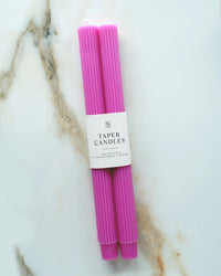 Two fluted, hand-dyed pink Wild Orchid Taper Candles by BOTANICA are bundled with a white label reading "Taper Candles" and rest on a white marble surface streaked with beige and brown veining.