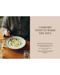 A person in a brown sweater enjoys creamy Nordic food at a wooden table. Next to them, the "Cookbook: Crumbs of Joy" by Cozy Publishing features “Comfort Food to Warm the Soul” and a quote on classic Finnish comfort recipes.