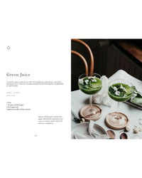 Two glasses of green juice topped with white edible flowers sit on a white tablecloth beside a wooden chair. Breakfast recipes from "Wonderful Morning - Recipes For A Delicious Start To the Day" by Cozy Publishing are shown on the left.