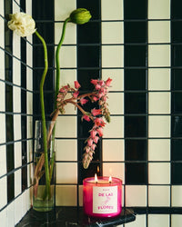 A ROEN De Las Flores Candle, crafted from coconut soy wax, burns on black marble next to a tall vase with two long-stemmed flowers, capturing the lively spirit of a Mexico City flower market against a black and white tiled wall.